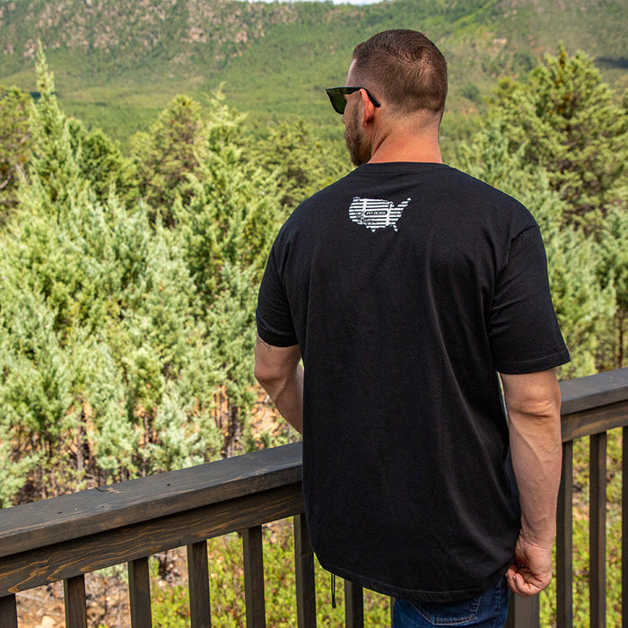Black Pit Boss Grills American Bull Men’s T-Shirt with USA graphic, worn outdoors overlooking forested landscape.