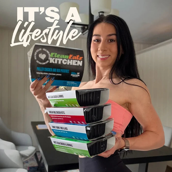 Woman smiling beside a Clean Eatz Kitchen box topped with stacked prepared meals like Arnold Bowl and Sweet-N-Sour Chicken
