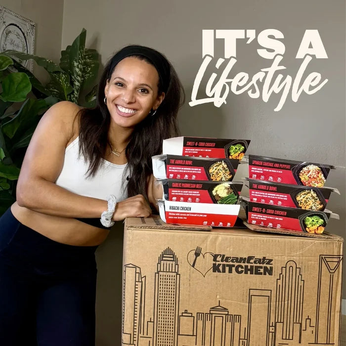 A smiling woman in a white sports bra and black leggings leans on a large cardboard box printed with the Clean Eatz Kitchen logo, with several Clean Eatz meal kit boxes stacked on top. Large white text in the upper right reads 'IT'S A Lifestyle'.