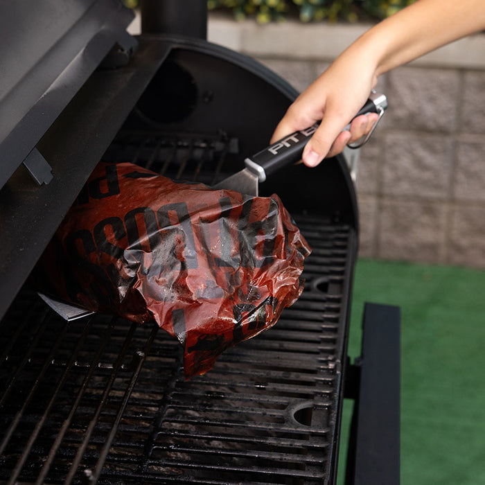 Pit Boss Big Head BBQ Spatula, silver with black handle and grey pit boss logo, lifting brisket wrapped in butcher paper from grill