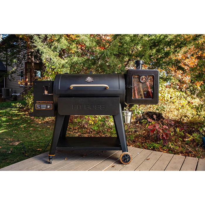 Grill on deck outside with trees and smoker sitting on side of grill. Pit boss Sportsman grill with orange accents.